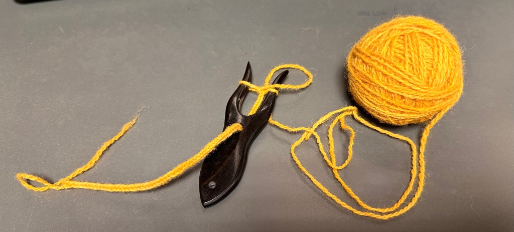 A lucet fork is sitting on a table next to a yellow ball of yarn. There is a lucet cord being made on the tines of the fork.